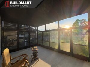 Interior view of a black framed glass porch enclosure with large panels, backyard view, seating area, and sunset light